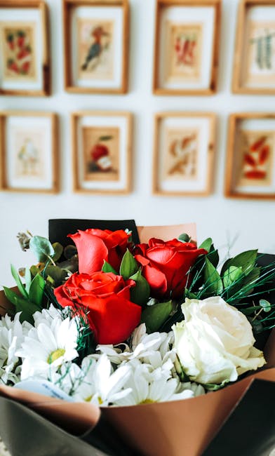 Close-up of a floral bouquet featuring vibrant red roses and white flowers, arranged with green foliage. The bouquet is wrapped in pink and beige paper with a black paper edge, held in hands in front 